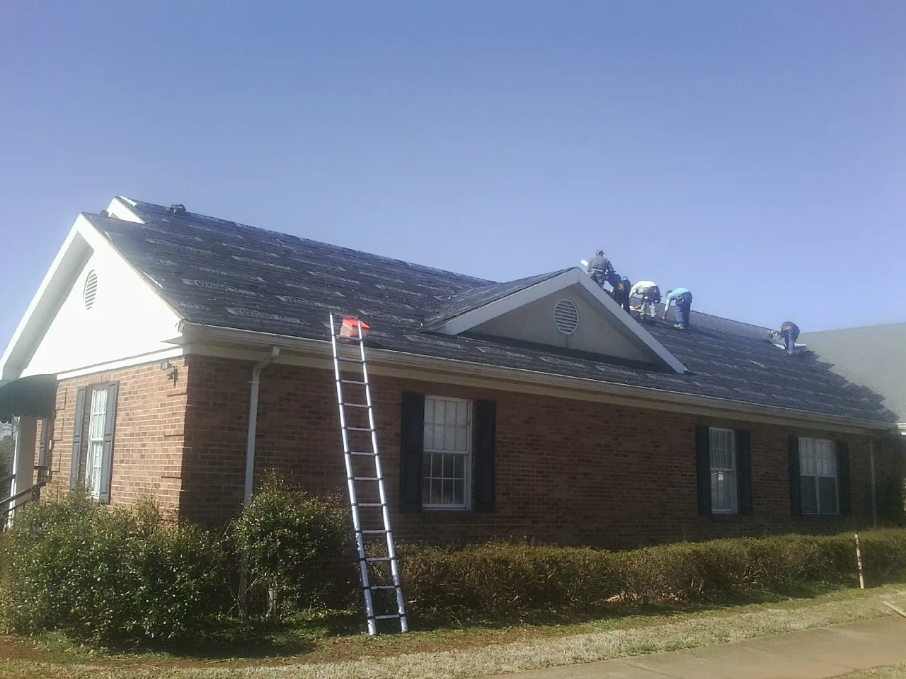 Crew tearing shingles off a brick ranch roof with a ladder set — Emergency Roof Repair work by our crew in Stroh